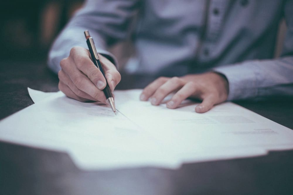 Two hands at a desk, one signing a printed document with a pen.